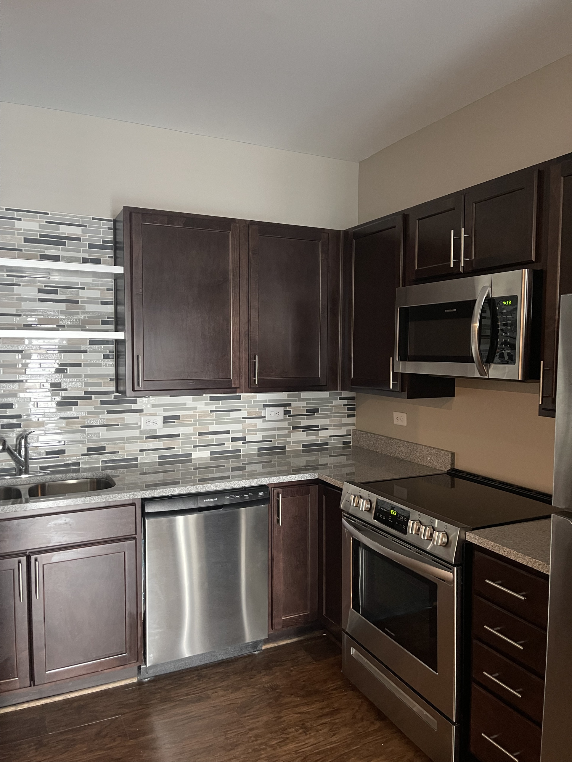 A kitchen corner with a mosaic tile backsplash, dark cabinetry, and a stainless steel microwave and range